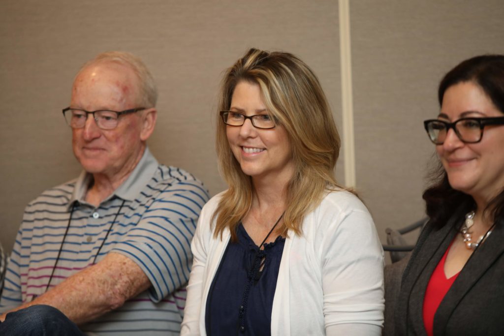 woman with blonde hair and white shirt smiling in between an older gentleman on the left side and a woman with dark hair and glasses on the right