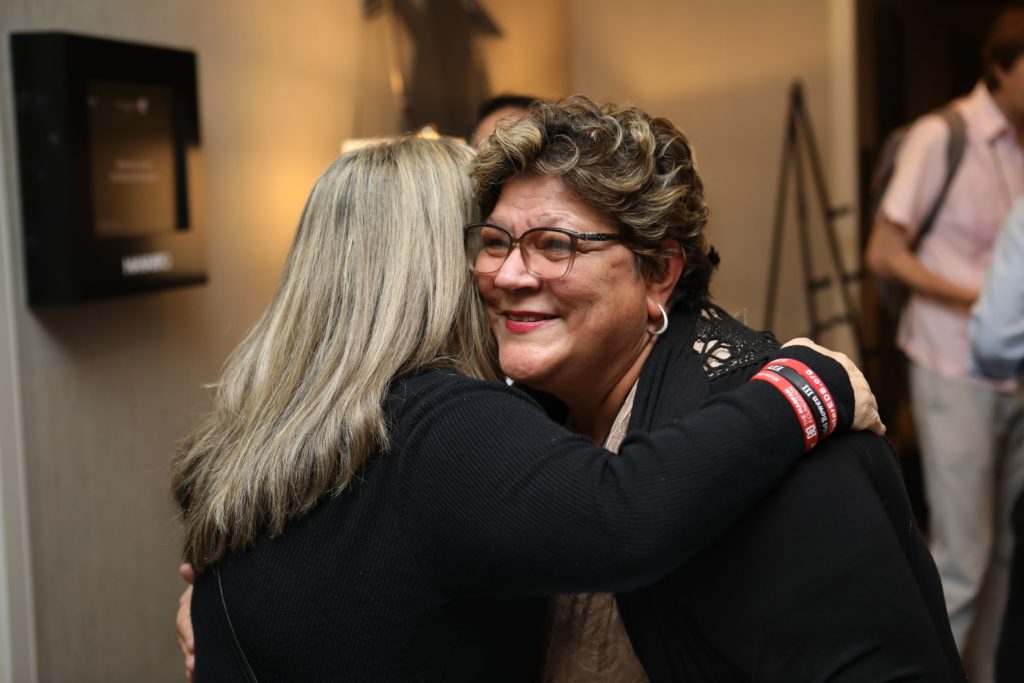 woman with blonde hair hugging another woman with dark, short, curly hair. Woman with dark hair is smiling