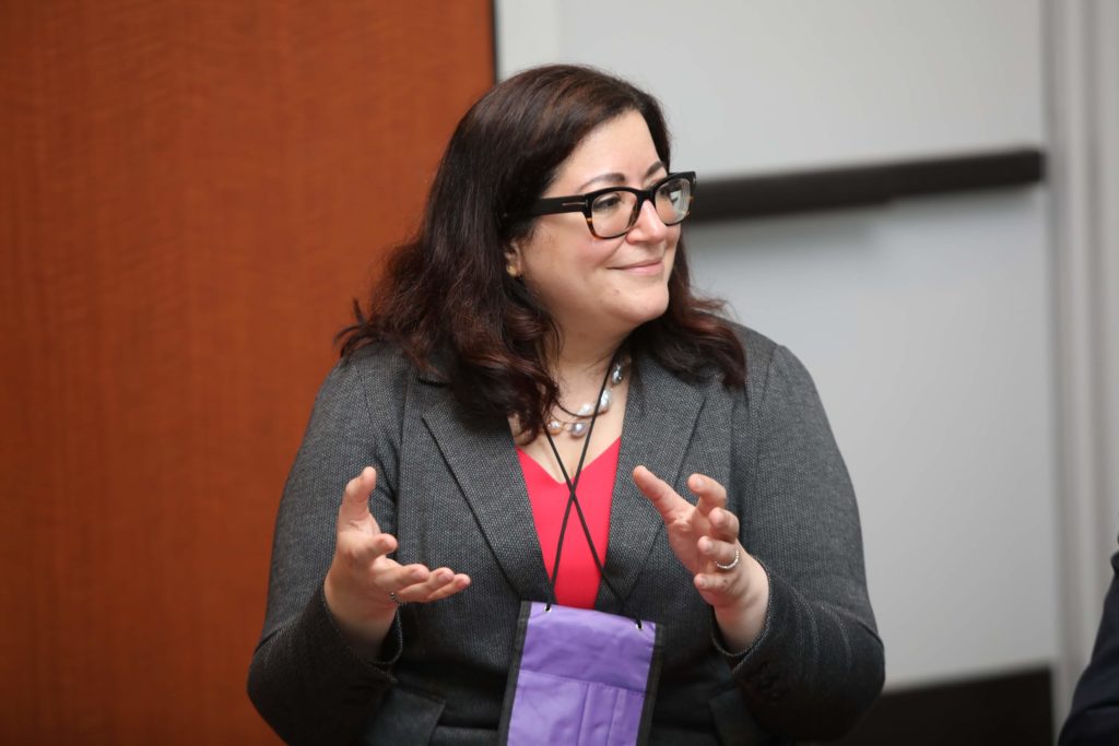 woman in gray blazer, pink blouse, dark hair, glasses, explaining something and smiling