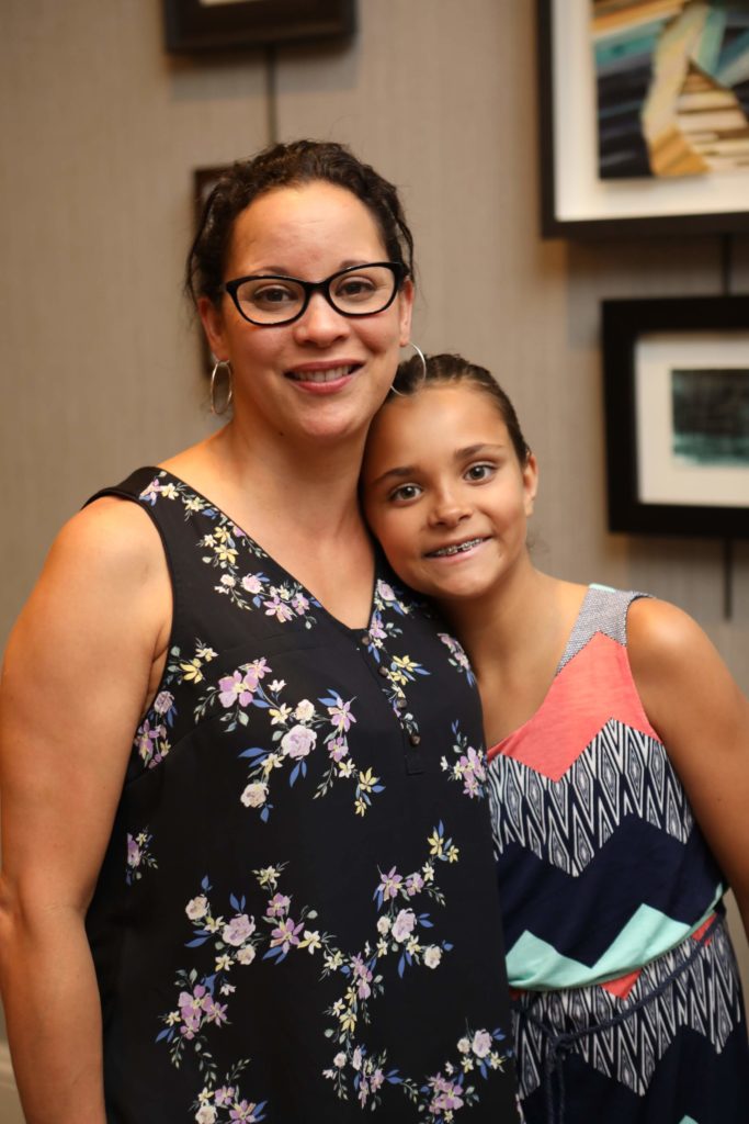 Woman with dark hair, dark shirt, glasses, standing next to and smiling with her daughter, who also has dark hair