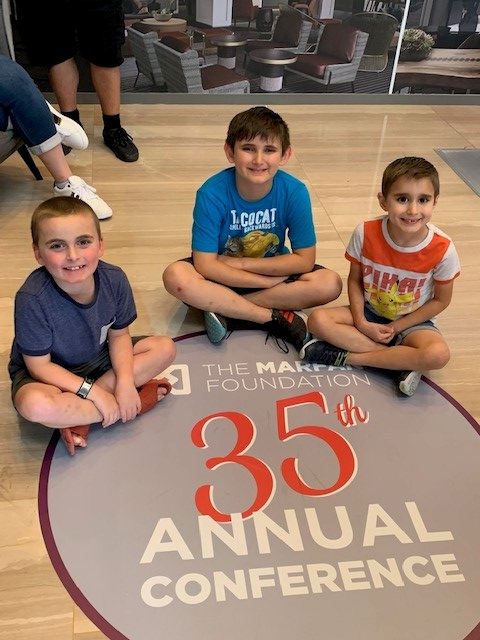 Three young boys with VEDS sitting cross legged on the floor smiling next to a floor sticker that says The Marfan Foundation 35th Annual Conference