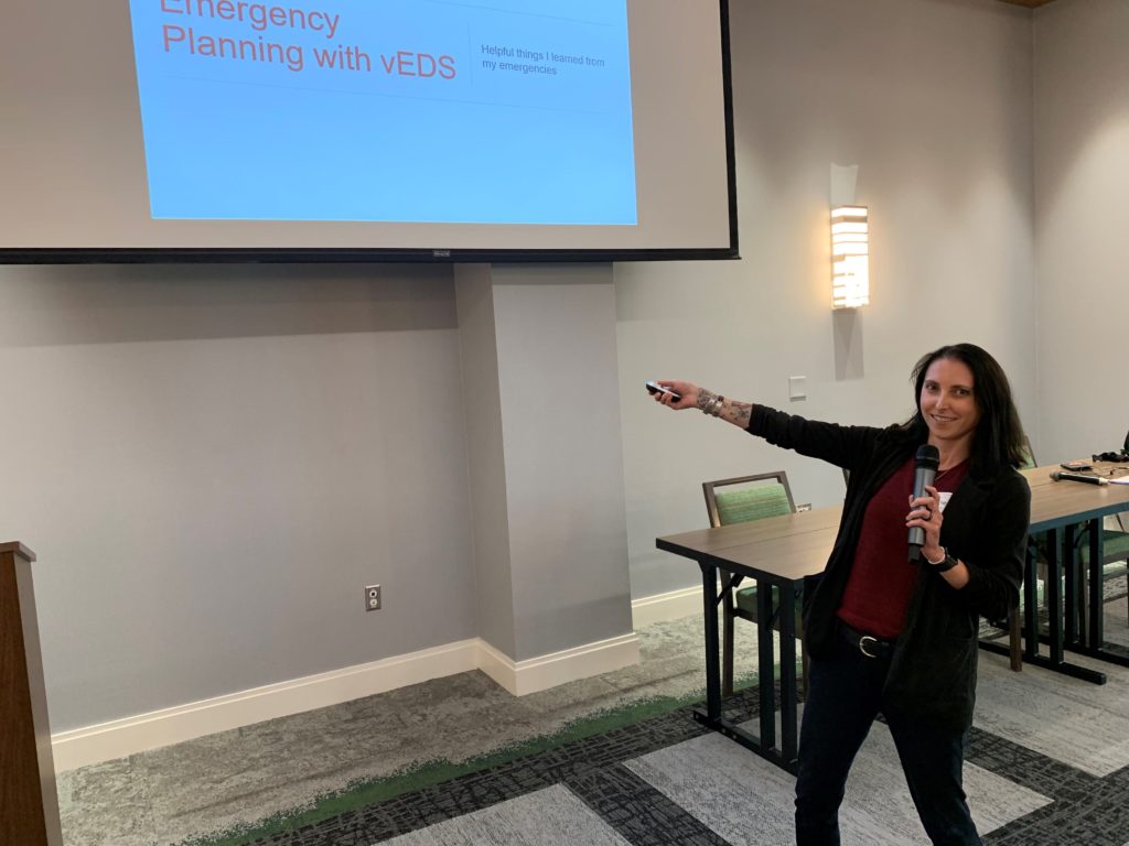 Woman with black hair and red shirt and black jacket pointing at powerpoint presentation titled Emergency Planning with vEDS