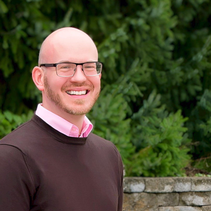 headshot image of Ryan smiling in front of green foliage