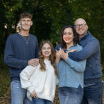 The Rodarmer family smiling for a portrait outdoors. A man, woman, teenage boy, and younger girl are posed together in front of green trees.