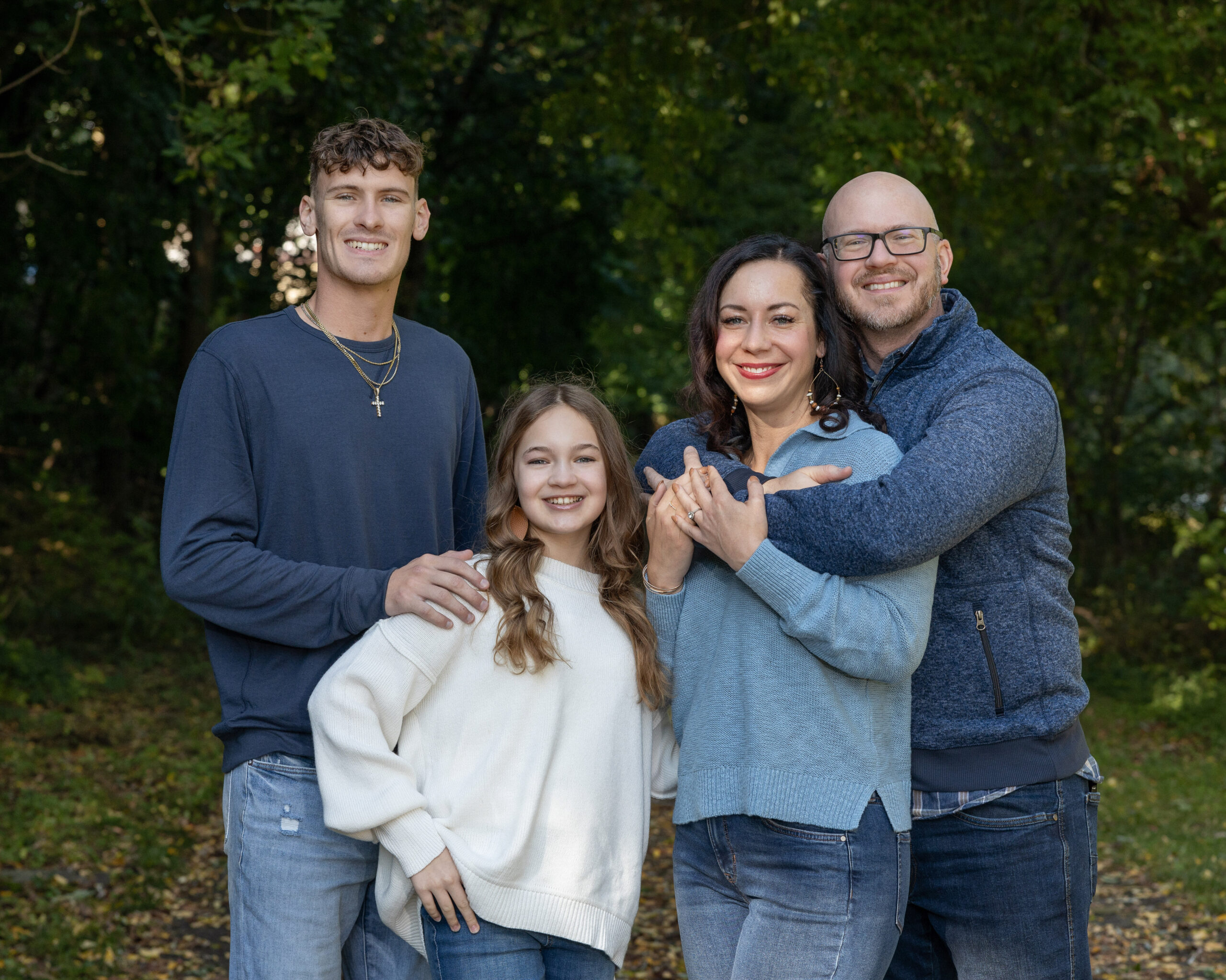 The Rodarmer family smiling for a portrait outdoors. A man, woman, teenage boy, and younger girl are posed together in front of green trees.