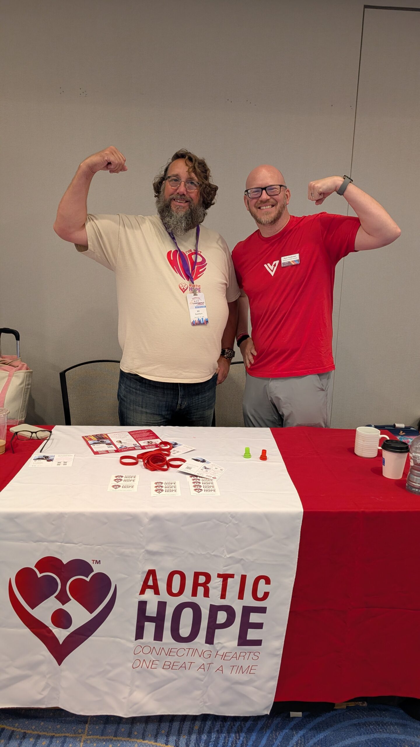 Ryan Rodarmer and Bill Barnhart flexing their biceps and smiling behind a table with an "Aortic Hope" banner at Conference.