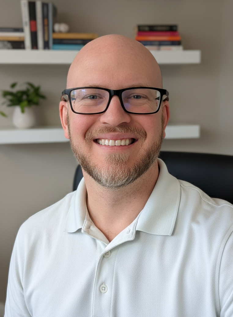 A headshot of Ryan Rodarmer, a smiling man with glasses and a beard, wearing a light-colored polo shirt in an office setting.